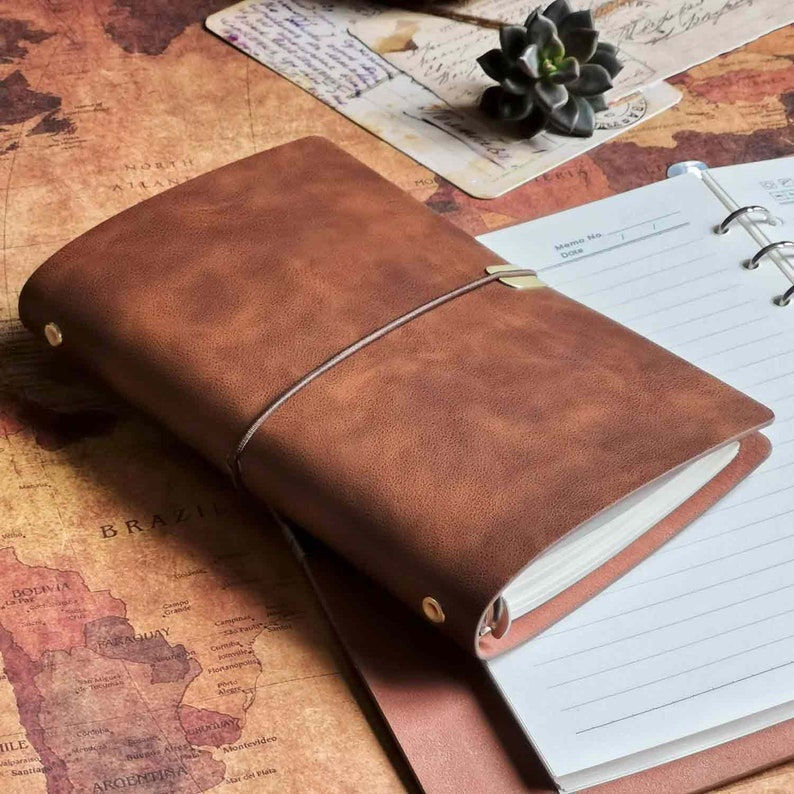 May include: A brown leather bound journal with a gold clasp and a ribbon closure. The journal is open to a blank page with lined paper.