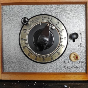 May include: A vintage timer with a black dial and a white face. The timer has a large knob and a smaller knob on the right side. The dial is marked with numbers from 0 to 60 and the words "EB(K) Technische Werkstätten Dresden". The smaller knob is marked with "1 s" and "1/10 s". The timer has a wooden frame and a gray background.