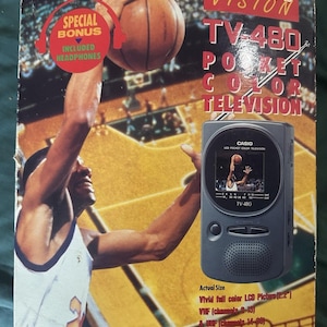 Pode incluir: Uma caixa de televisão a cores de bolso Casio Sports Vision TV-480 com um gráfico de jogador de basquetebol. A caixa exibe o nome do produto, uma especificação de ecrã LCD de 2,2 polegadas e um emblema circular vermelho com auscultadores.
