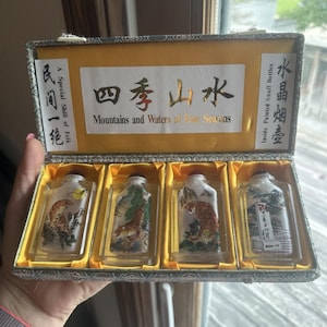 May include: A rectangular presentation box containing four clear glass snuff bottles with inside-painted tiger and landscape imagery. The box features a yellow interior lining and a white label with the text Mountains and Waters of Four Seasons.
