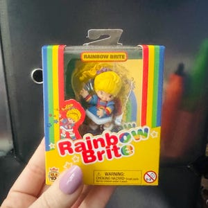 May include: A Rainbow Brite collectible figurine inside a rectangular cardboard box with a clear plastic window. The packaging features a yellow background, rainbow-colored side borders, and the text Rainbow Brite in red and green letters.