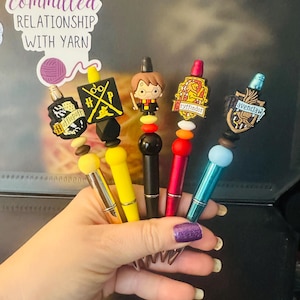 Peut inclure: Cinq stylos avec des dessus décoratifs aux motifs variés. Les stylos ont des corps de couleurs différentes, dont jaune, noir et rouge. Les dessus comprennent un personnage de dessin animé et des blasons de maisons. L'image comprend également un autocollant qui dit "IN A committed RELATIONSHIP WITH YARN."