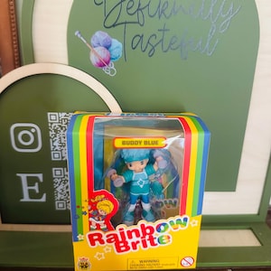 May include: A Buddy Blue Rainbow Brite action figure in its original rectangular packaging. The box features a rainbow border, the Rainbow Brite logo, and a clear plastic window displaying the blue-haired character holding small dumbbells.
