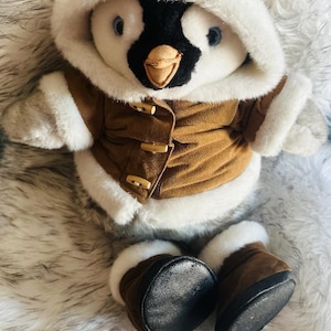 May include: A plush penguin toy wearing a brown hooded coat with white faux fur trim and three wooden toggle buttons. The penguin has a black and white face, a tan beak, and is wearing matching brown boots with white faux fur cuffs.