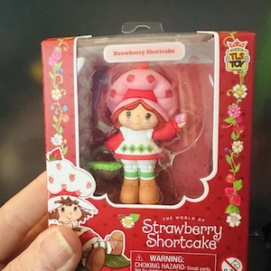 May include: A Strawberry Shortcake collectible figure in a red box. The figure wears a pink strawberry-patterned hat, a white dress with green diamond accents, and brown boots. The packaging features the text The World of Strawberry Shortcake.