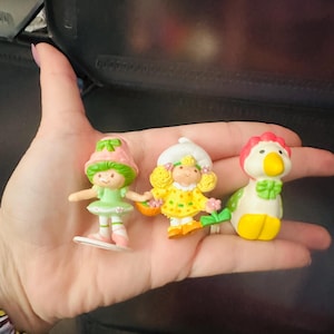 May include: Three small plastic figurines including a character with a pink hat and green dress, a character with a white hat and yellow polka dot dress, and a white bird figurine with a yellow beak and red crest, all standing on a palm.