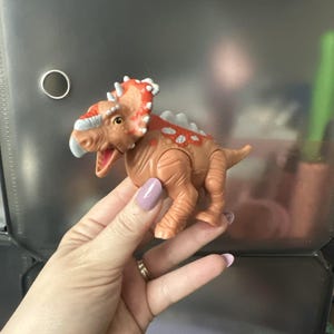 May include: A tan plastic dinosaur figurine with a frilled head, white spikes along the back, and orange markings on the head and spine. The dinosaur has a light gray beak, a wide open mouth showing a pink interior, and a textured skin surface.