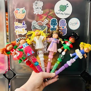 May include: A selection of decorative pens with doll figures as toppers. Each pen has a unique design with colourful beads and a doll figure. The dolls are dressed in various outfits, adding a playful touch to the writing instruments.