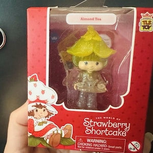 May include: A collectible Almond Tea figurine from The World of Strawberry Shortcake inside a red cardboard box. The translucent, glittery character wears a yellow flower hat and holds a small tray. The packaging features Strawberry Shortcake.