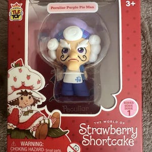 May include: A Peculiar Purple Pie Man collectible figure from The World of Strawberry Shortcake series 1, encased in a red rectangular box with a clear plastic window. The figure features a white mustache, purple hat, and blue outfit.