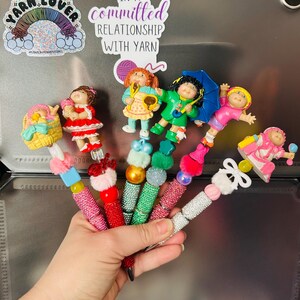 Könnte beinhalten: Eine Sammlung von fünf dekorativen Stiften mit Puppenaufsätzen im Cabbage Patch Kids-Stil. Jeder Stift hat ein einzigartiges Design mit bunten Perlen, Strasssteinen und flauschigen Pompons. Die Puppenfiguren tragen verschiedene Outfits.