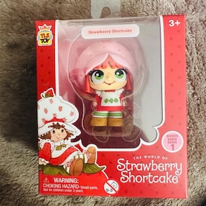 May include: A Strawberry Shortcake collectible figure in a red box with a clear plastic window. The figure has a pink hat, red hair, green eyes, and a white apron over a red dress. The packaging features the text The World of Strawberry Shortcake.