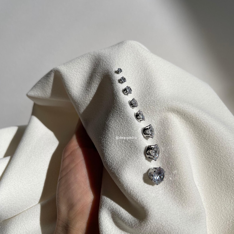 Row of cubic zirconia stud earrings in various sizes (3mm-10mm), displayed on soft white fabric, crafted with 316L surgical steel, hypoallergenic and designed for sensitive ears