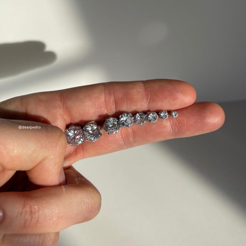 Close-up of cubic zirconia stud earrings in sizes 3mm to 10mm displayed on fingers, crafted from 316L surgical steel for hypoallergenic wear with a brilliant diamond-like sparkle