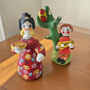 May include: Two painted clay figurines. One figure has black hair, yellow earrings, and a red dress with colorful floral patterns, holding a small yellow plate. The second figure has brown hair, a red outfit, and holds a yellow stringed instrument.