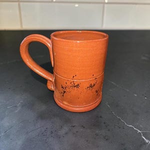 May include: A terracotta-colored ceramic mug with a smooth, cylindrical body and a curved handle. The lower section of the mug features a horizontal embossed line and scattered dark brown, speckled markings across the surface of the clay.