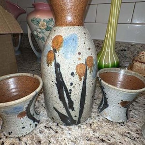 May include: A ceramic sake set consisting of one tall bottle and two matching cups. The pieces feature a speckled cream base with brown rims, black painted floral stems, and orange flower heads, accented by patches of light blue glaze.
