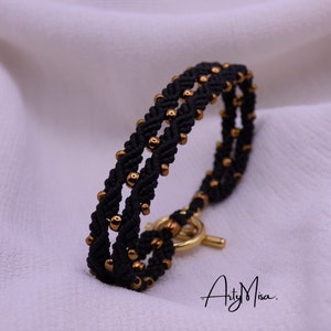 May include: A black braided bracelet with gold beads and a gold clasp. The bracelet is on a white background.