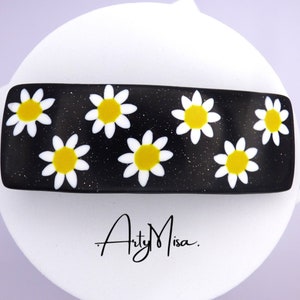 Könnte beinhalten: Schwarzer Haarklammer mit weißen Gänseblümchen und gelben Zentren auf einem glitzernden schwarzen Hintergrund. Die Klammer hat den Text "Arty Misa." unten aufgedruckt.