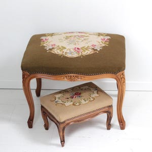 May include: A large olive green upholstered footstool with a floral needlepoint center and carved wooden legs, paired with a smaller beige upholstered footstool featuring a matching floral needlepoint design and carved wooden legs.
