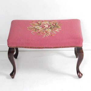 Puede incluir: Un otomán vintage con una parte superior tapizada de color rosa empolvado. La parte superior presenta un diseño floral de punto de cruz en tonos rosas, verdes y crema. Las patas curvas son de color marrón oscuro. Los bordes de la parte superior están adornados con pequeñas tachuelas de latón.