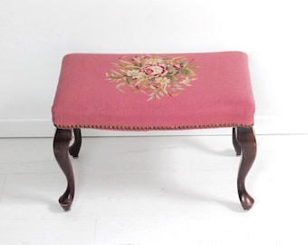 May include: A vintage footstool with a dusty rose upholstered top. The top has a floral needlepoint design in pinks, greens, and cream. The curved legs are dark brown. The top's edges are trimmed with small brass tacks. The stool measures approximately 76 cm long.