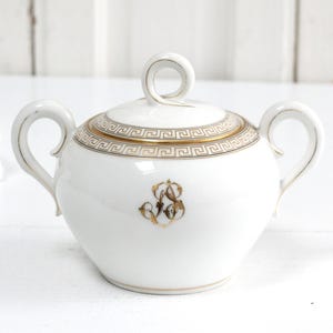 May include: White porcelain sugar bowl with a lid, featuring gold Greek key pattern around the rim and a gold monogram. The bowl has two curved handles and a loop handle on the lid. A classic piece for serving sugar.