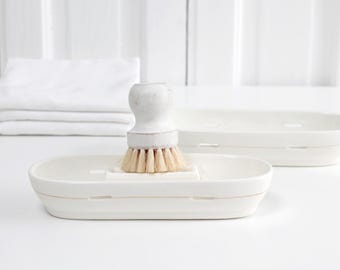May include: A white ceramic dish brush with a light wood handle and natural bristles sits in a matching white soap dish. A second dish and folded white towels are also visible.