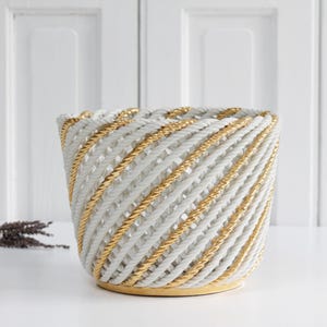 May include: A white and gold rope basket with a woven design. The basket is round and has a gold base.