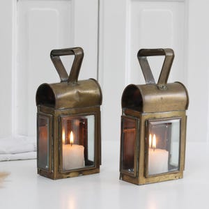 May include: Two antique brass lanterns with glass panels and a handle. Each lantern holds a lit white candle, casting a warm glow. The lanterns are placed on a white surface, creating a cozy, vintage aesthetic.