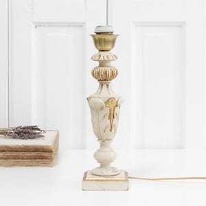 May include: A cream-coloured vintage table lamp with gold detailing. The lamp features an ornate design, a square base, and a brass-coloured top. A stack of old books and dried lavender are in the background.