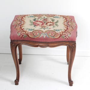 Puede incluir: Un pequeño taburete antiguo con una parte superior de tapiz floral en tonos rosa, crema y verde. El marco de madera presenta patas curvas y tallas decorativas, mostrando una estética vintage.