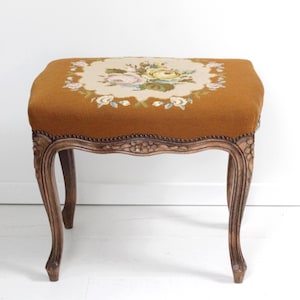 Puede incluir: Un taburete antiguo con una parte superior bordada floral. El asiento es de color marrón dorado cálido, y el bordado presenta un ramo de flores rosas, amarillas y blancas. Las patas son de madera tallada con un acabado marrón oscuro.