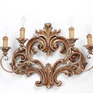 May include: A wall sconce featuring a central carved wooden frame with ornate scrollwork and a distressed gold finish. The fixture holds four light sockets with beige candle covers and clear flame-shaped bulbs extending from curved metal arms.