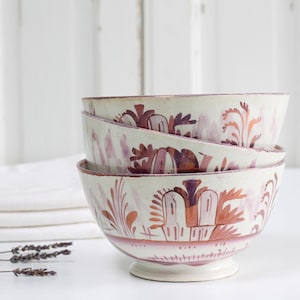 Puede incluir: Tres cuencos de cerámica apilados, con base crema y diseño floral burdeos y naranja. Los cuencos están sobre un fondo blanco, con un paño blanco doblado cerca. Los cuencos miden aproximadamente 15-20 cm de diámetro.