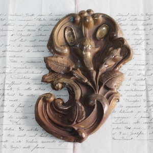 May include: A decorative, ornate, wooden floral design with a brown and gold finish. The design is intricate and detailed, with swirling leaves and scrolls.