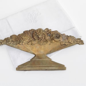 Puede incluir: Centro de mesa floral de metal en tono bronce vintage. La pieza decorativa presenta una disposición detallada de flores y follaje esculpidos. Descansa sobre una base de pedestal, sobre un fondo blanco con una servilleta de lino blanca.