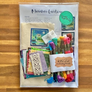 May include: A craft kit titled Miniature Landscape containing a printed instruction sheet, a square of off-white fabric with a small landscape embroidery, assorted colourful fabric scraps, a piece of white lace, and a bundle of embroidery threads.