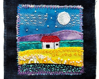 May include: A textile art piece depicting a night scene with a white house, a full moon, and stars. The artwork features a red-roofed house, a yellow field, and a blue sky with white lace and embroidered details. The piece is framed with a dark blue border.