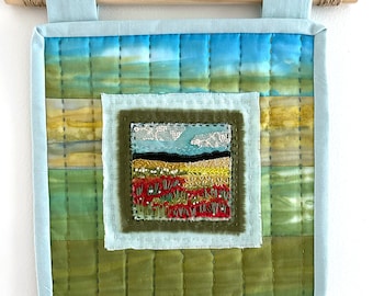 May include: A quilted wall hanging with a blue, green, and brown color scheme. The center of the piece features a small embroidered landscape with a red, yellow, and green field.
