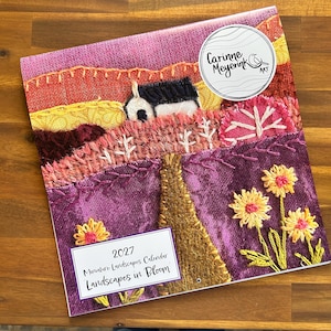May include: A 2027 wall calendar titled Miniature Landscapes Calendar Landscapes in Bloom featuring a textured fabric embroidery of a landscape with a house, hills, and flowers in shades of purple, orange, and yellow with a circular logo.