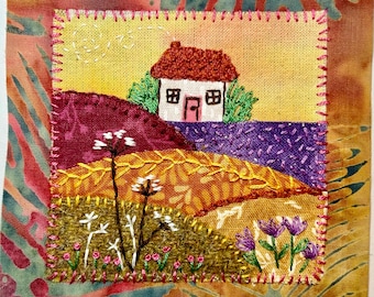 May include: Hand-stitched fabric art featuring a quaint house with a red roof and a pink door, set against a yellow and orange sky. The landscape includes purple and green hills, with embroidered flowers and plants in the foreground. The artwork is framed with a decorative border.