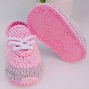 May include: Pink crocheted baby shoes with gray accents and white laces. The soles are pink and have a cartoon design of a sun, moon, birds, and a giraffe.