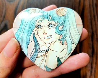 May include: Heart-shaped button featuring a smiling cartoon mermaid with blue hair and a seashell hair accessory. The mermaid is wearing a pearl bracelet. The button is held in a hand.