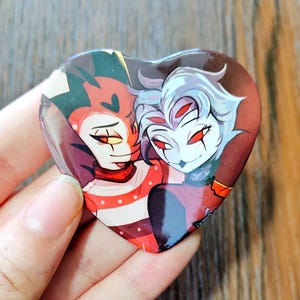 May include: Heart-shaped button featuring two cartoon characters with red and white accents. One character has red skin and a black and yellow headpiece. The other has pale skin and white hair. The button is held by a person.