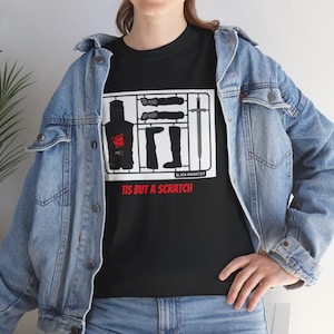 Peut inclure: T-shirt noir avec un motif de kit de Chevalier Noir, comprenant des pièces d'armure et une épée, et le texte "TIS BUT A SCRATCH" en rouge. Le t-shirt est porté avec une veste en jean bleue.