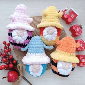 May include: Four handmade crochet gnomes in pink, yellow, blue, and orange. Each gnome has a white beard and a matching hat. Decorative red train and berries are also in the image. The gnomes are on a woven surface.