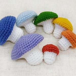 K&ouml;nnte beinhalten: Ein Set aus sieben geh&auml;kelten Pilzen in einem Regenbogen aus Farben. Die Pilze sind alle unterschiedlich gro&szlig; und haben wei&szlig;e Stiele.