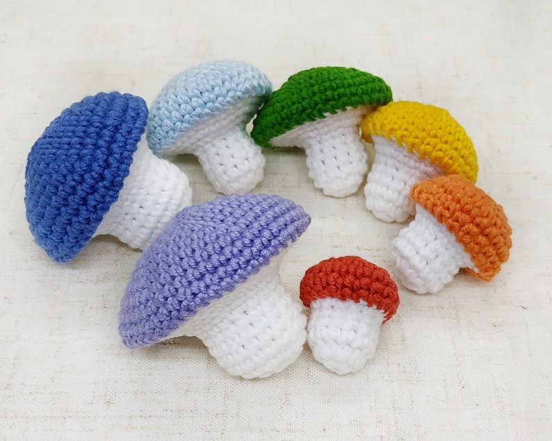 K&ouml;nnte beinhalten: Ein Set aus sieben geh&auml;kelten Pilzen in einem Regenbogen aus Farben. Die Pilze sind alle unterschiedlich gro&szlig; und haben wei&szlig;e Stiele.