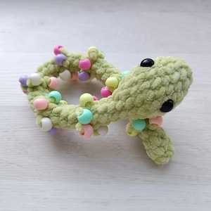May include: A handmade, light green crocheted lizard toy with black button eyes and colourful beads. The plush toy is decorated with pastel-coloured beads in pink, yellow, blue, and white. The lizard is lying on a white surface.